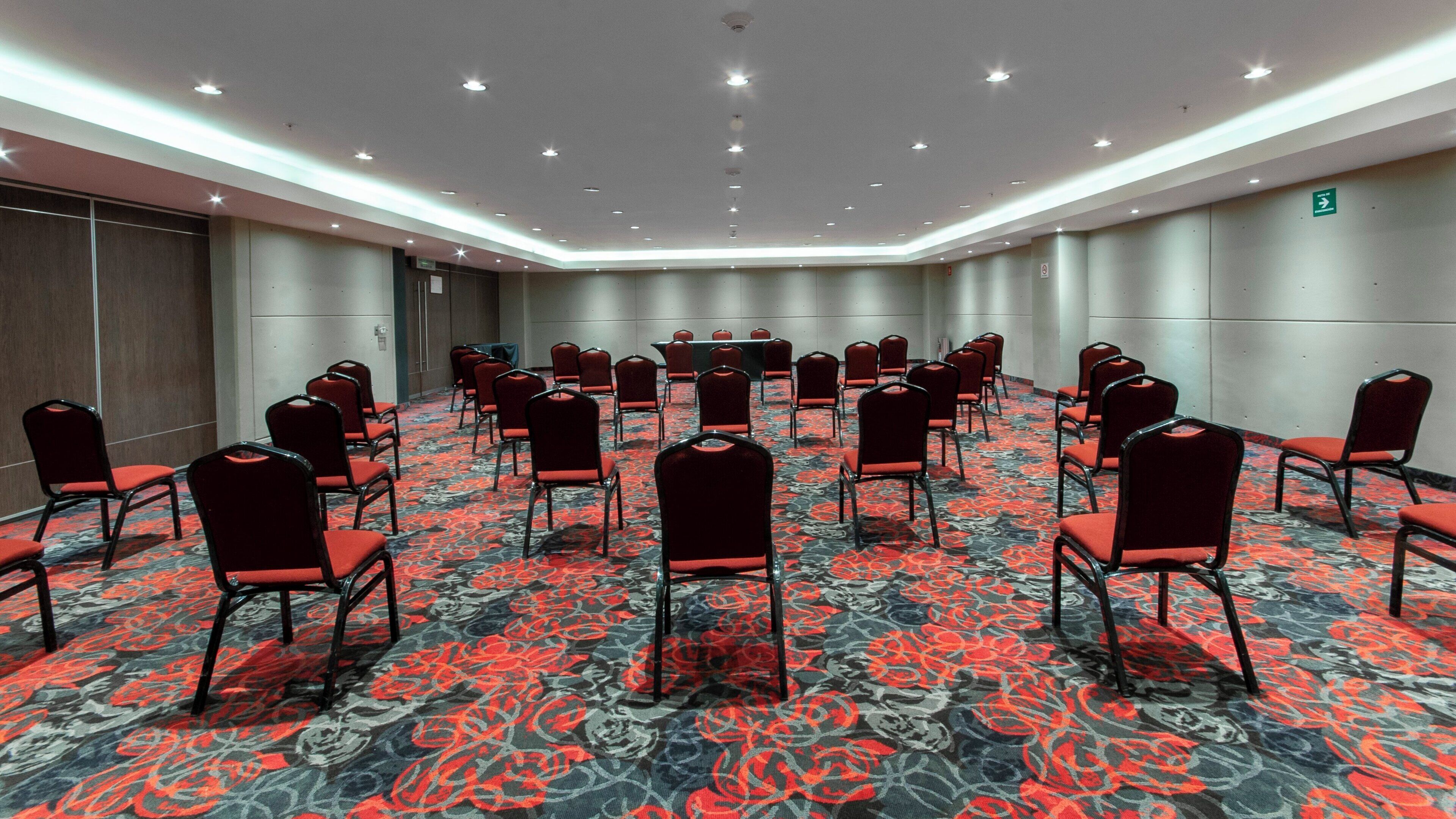 A conference room with rows of chairs facing a table, carpeted floor, and ceiling lights.