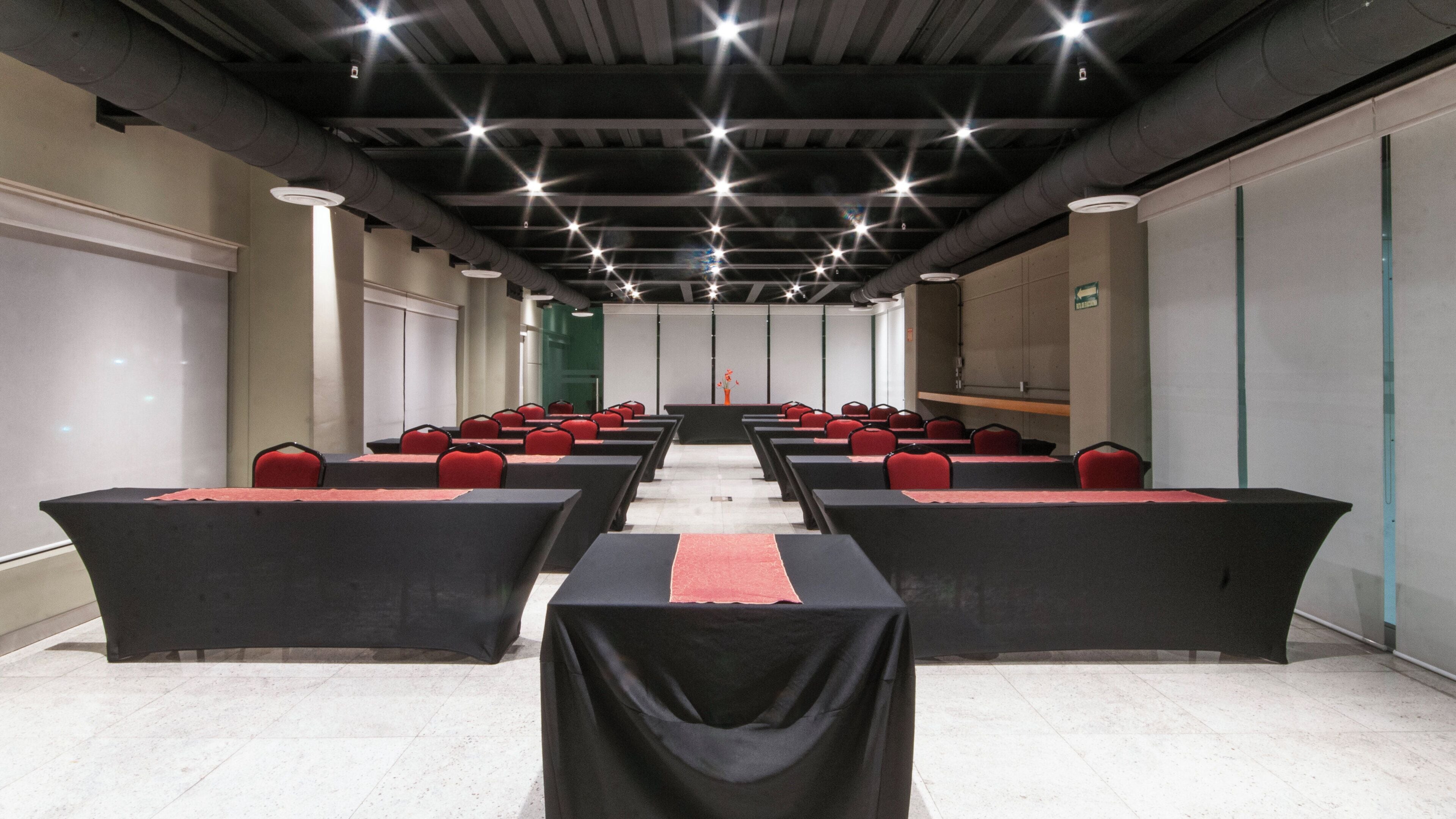 A conference room with rows of tables and chairs, a long table at the front, and a high ceiling with visible ductwork.