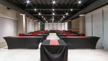 A conference room with rows of tables and chairs, a long table at the front, and a high ceiling with visible ductwork.