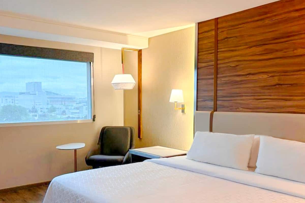 Habitación de hotel con una cama grande, un escritorio, una silla, un ventanal con vista a la ciudad y dos lámparas de pared.