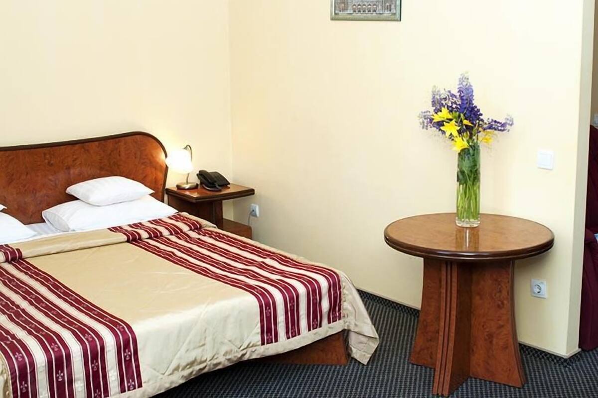 Habitación de hotel con cama, mesita de noche con lámpara y teléfono, una mesa redonda de madera con un jarrón de flores, y un cuadro en la pared.