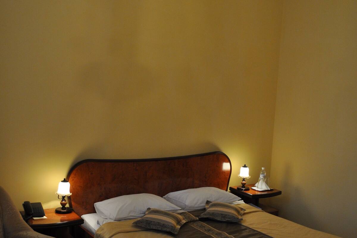 Habitación de hotel con cama doble, cabecero de madera, dos mesitas de noche con lámparas, una silla y una araña.