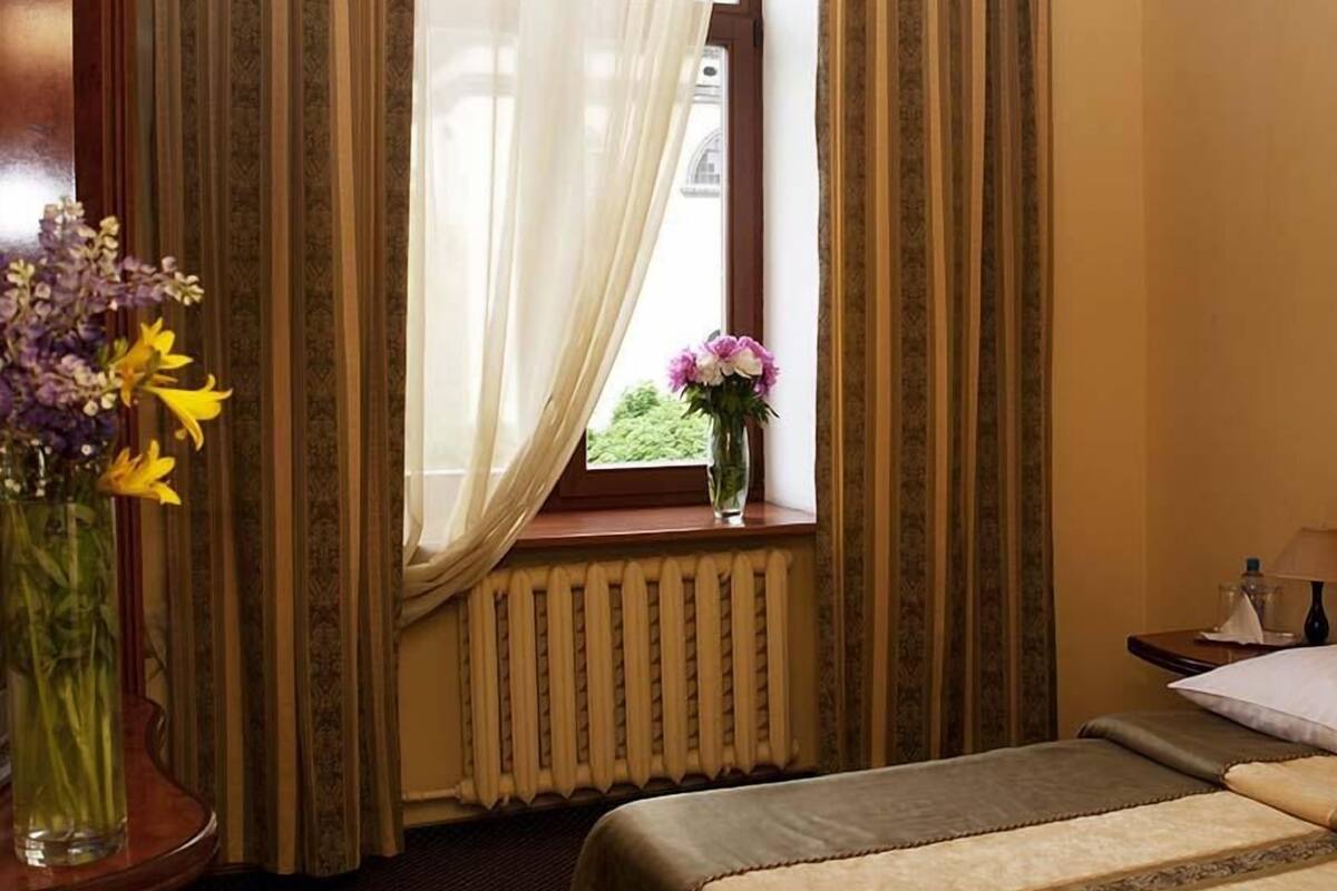 Una habitación de hotel con una cama, una ventana con cortinas, un florero con flores y un radiador.