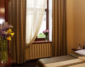 A hotel room with a bed, a window with curtains, a vase with flowers, and a radiator. - Wien Hotel (Lviv)