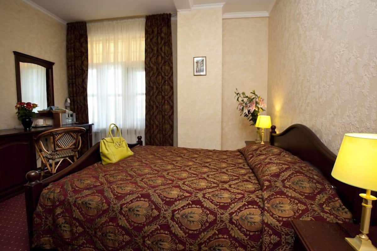 A hotel room with a large bed, a wooden nightstand with a lamp, a chair, a desk with a mirror, and a window with curtains.
