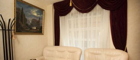 Una habitación con dos sofás de cuero color beige, una mesa de centro con revistas, un cuadro enmarco de un edificio histórico y una ventana con cortinas rojas.