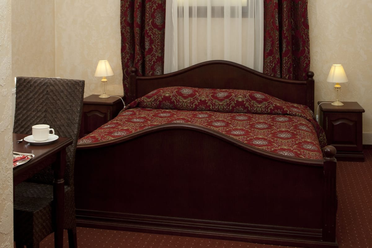 A hotel room with a wooden bed, a desk with a chair, two bedside lamps, and a window with sheer curtains and patterned drapes.
