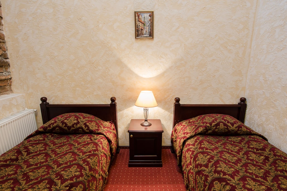 A room with two wooden beds, each with a red and gold patterned bedspread, a small bedside table with a lamp, and a framed picture on the wall.