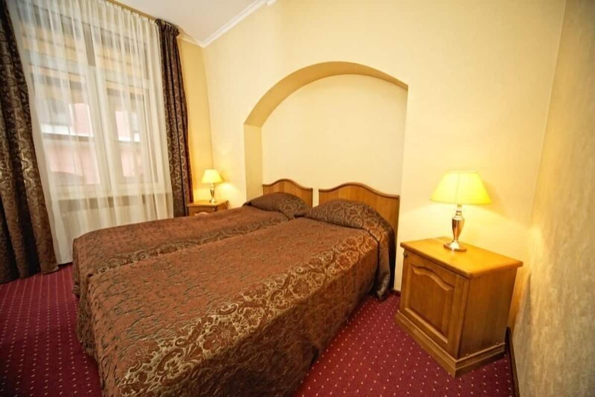 A double bed with a patterned bedspread, two bedside lamps, a wooden nightstand, and a window with sheer curtains.