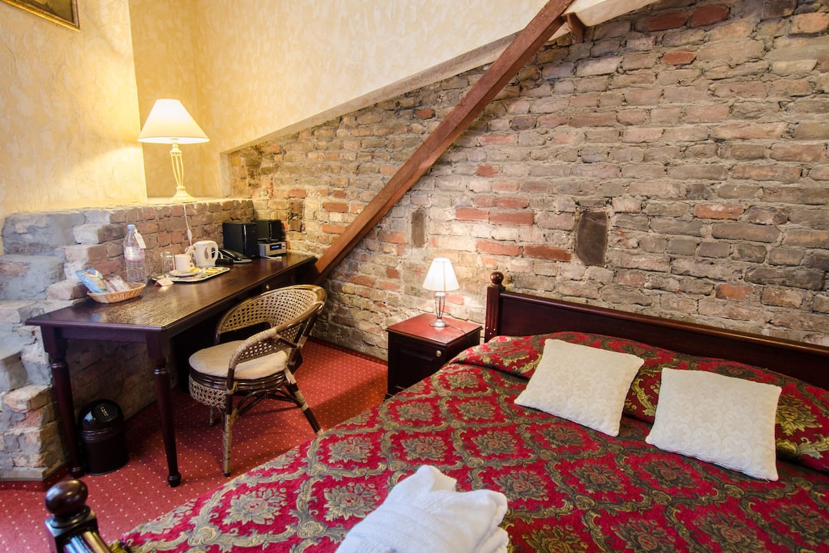 A cozy room with a brick wall, a wooden desk, a wicker chair, a bed with red and gold bedding, and a lamp.