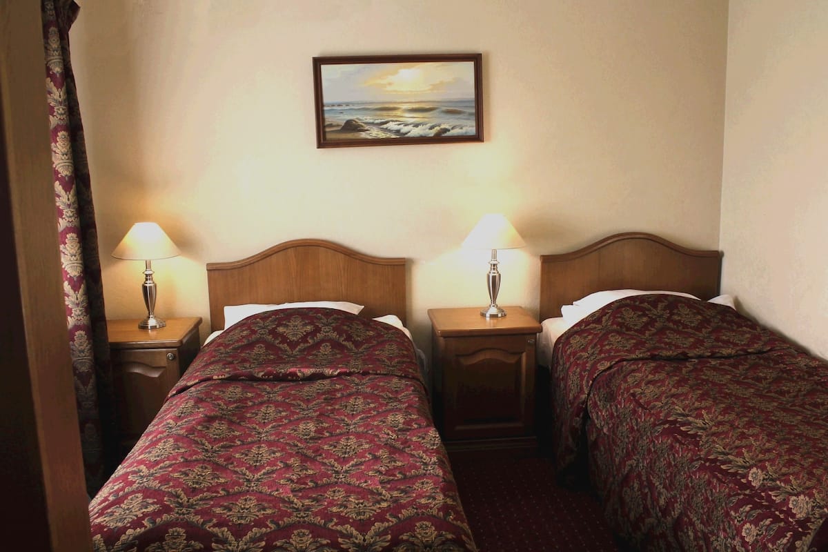 A hotel room with two single beds, each with a wooden headboard, a nightstand with a lamp, and a painting of a seascape on the wall.