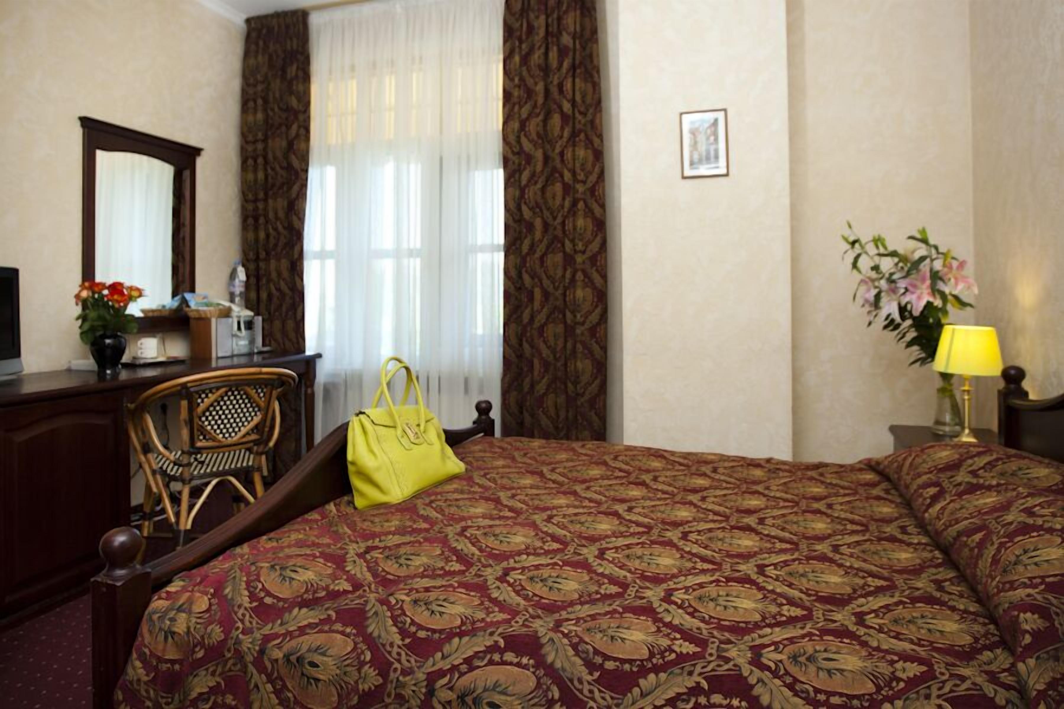 A hotel room with a large bed, a desk with a chair, a mirror, a lamp, and a vase with flowers.