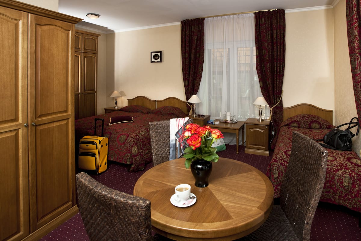 A hotel room with two beds, a wooden wardrobe, a round table with a vase of flowers, and a suitcase.