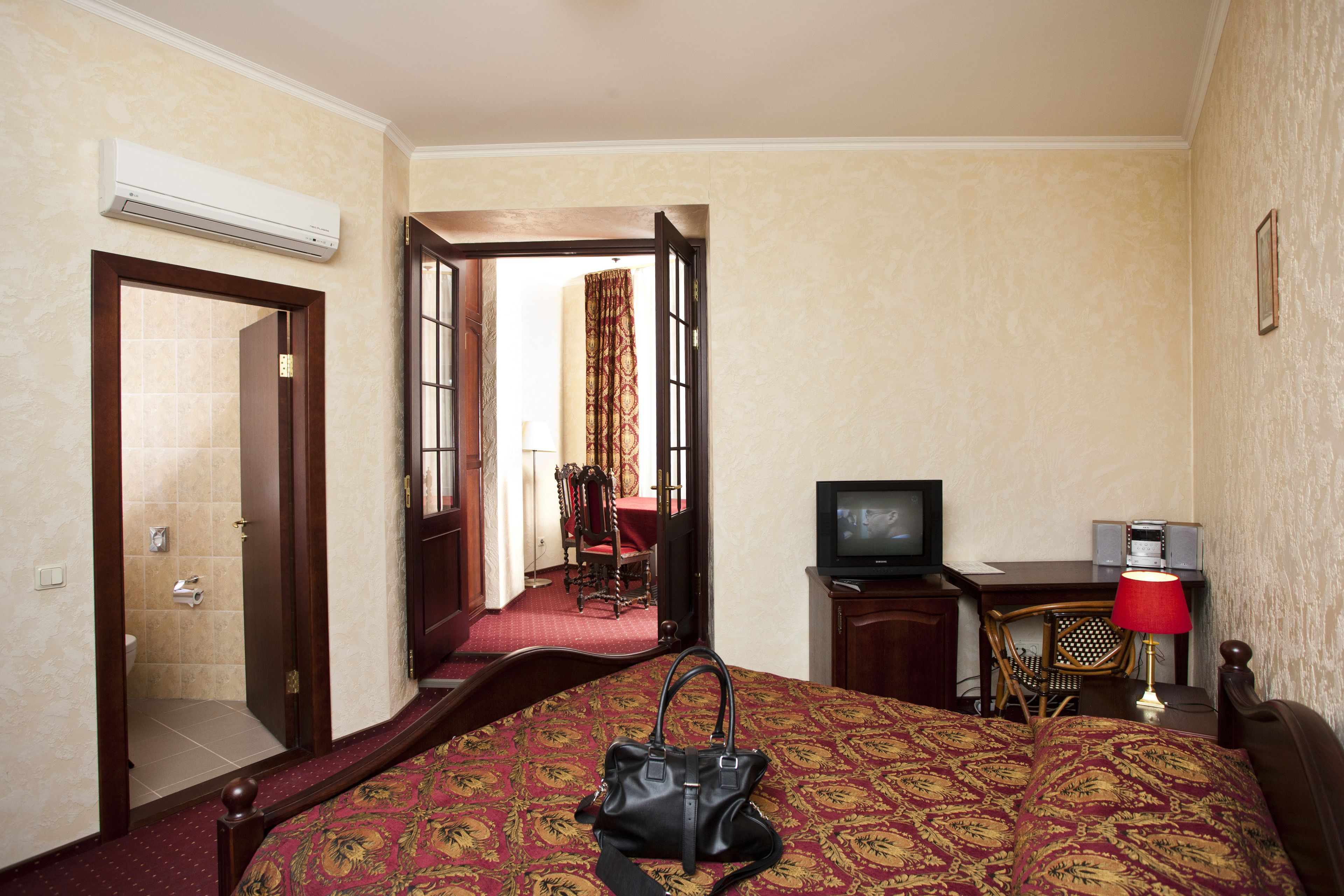 A hotel room with a bed, a television, a desk with a lamp, a chair, and an air conditioning unit.