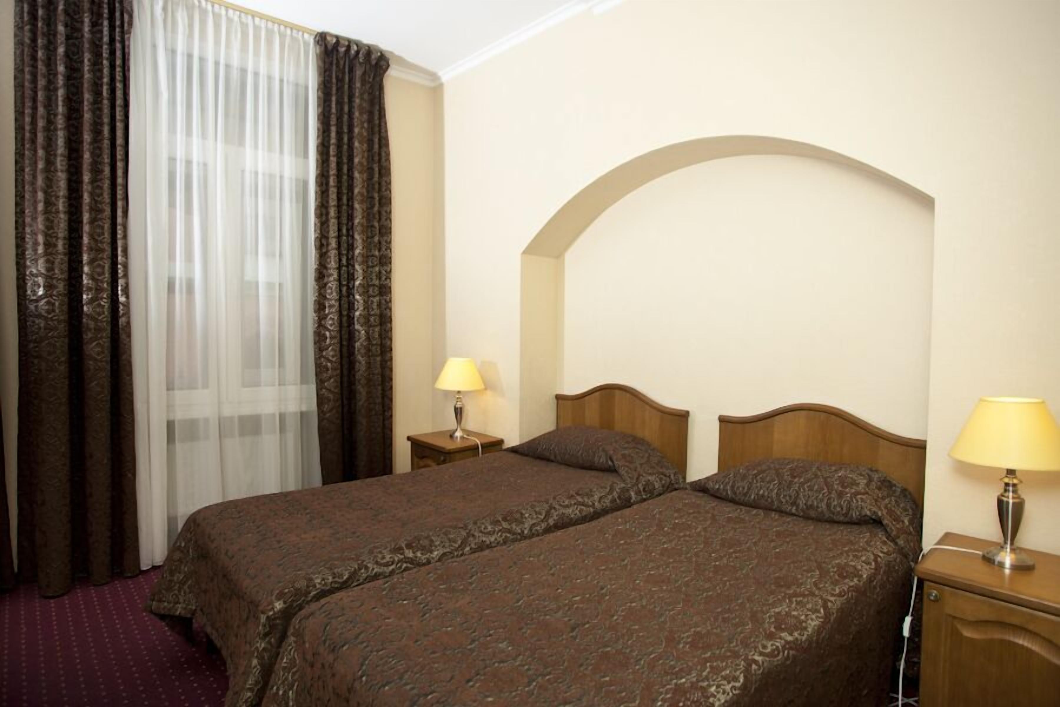 Une chambre d’hôtel avec deux lits, une tête de lit en bois, une petite table de chevet avec une lampe et une fenêtre avec des rideaux légers.