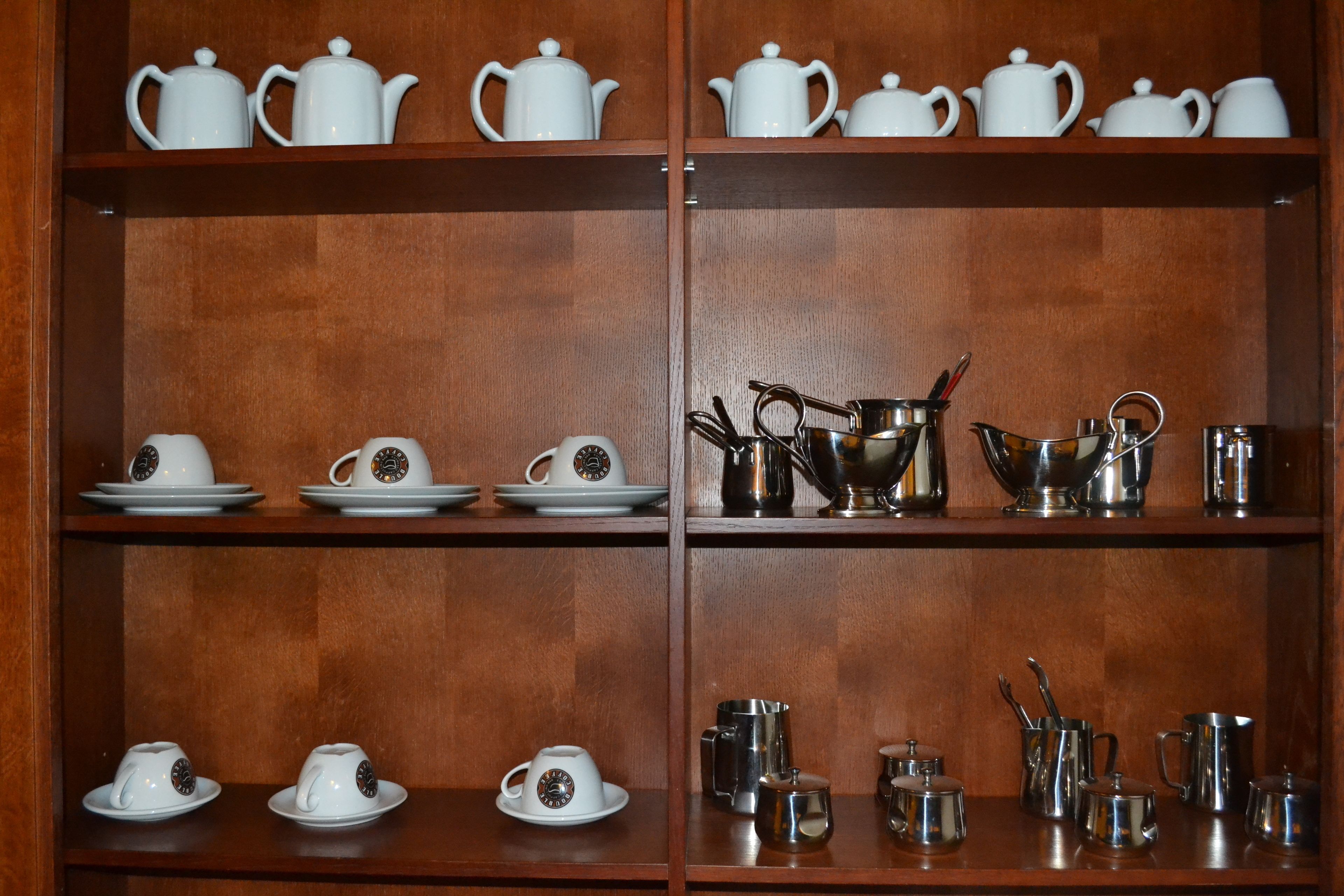 A wooden cabinet with shelves containing white teapots, cups, and silver teacups.