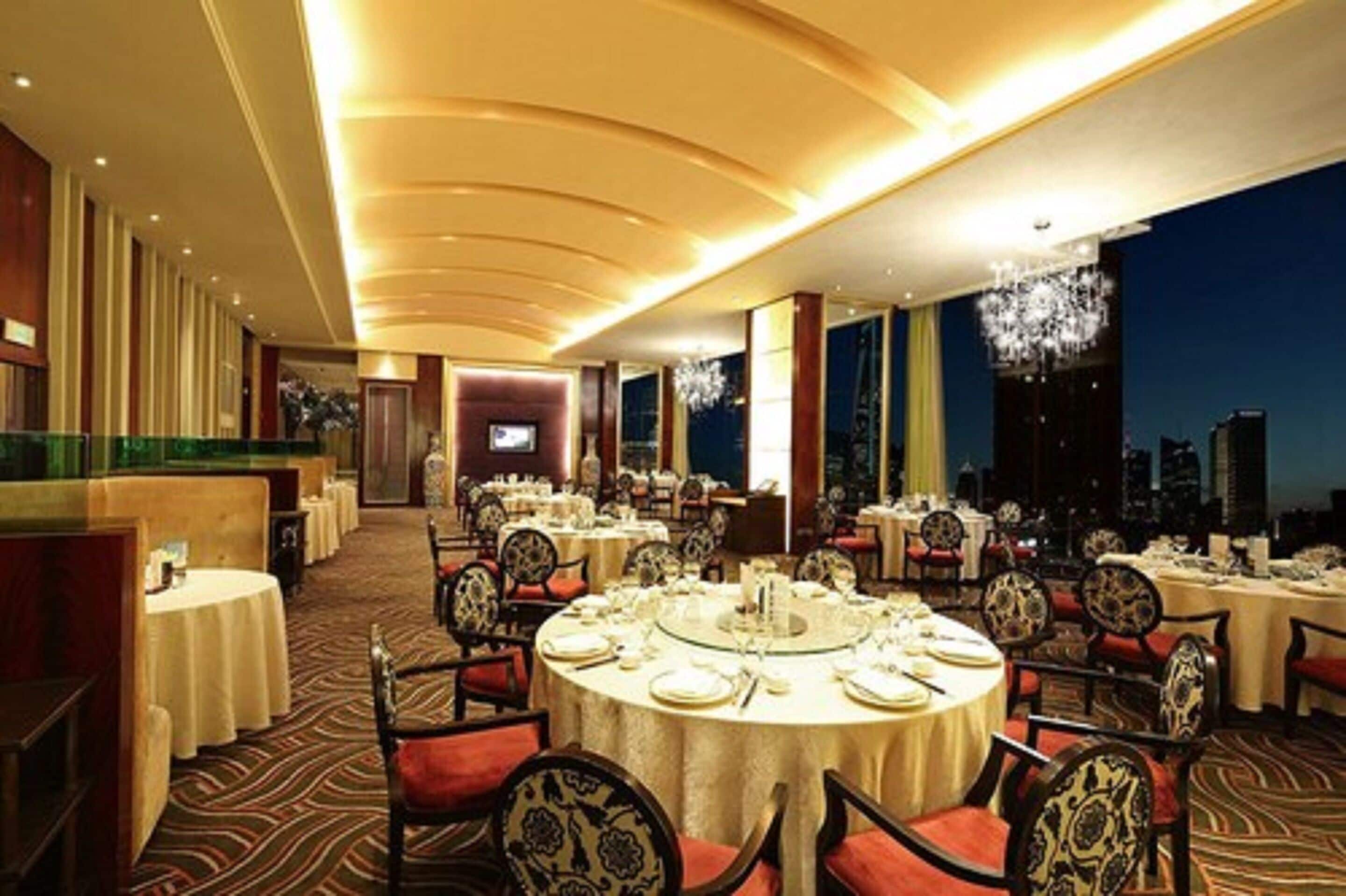 A spacious dining area with round tables and chairs, a city view, and a modern ceiling design.