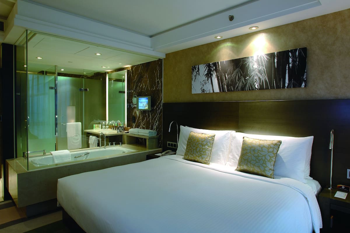 A hotel room with a large bed, a glass-enclosed shower, a vanity with a mirror, and a wall-mounted TV.