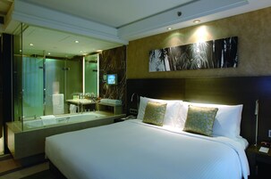 A hotel room with a large bed, a glass-enclosed shower, a vanity with a mirror, and a wall-mounted TV.