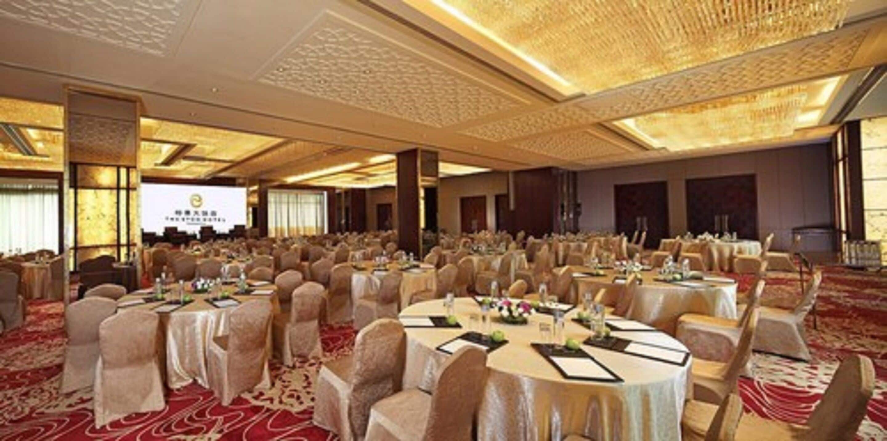 Un salón de banquetes con mesas redondas, sillas y un escenario para presentaciones.