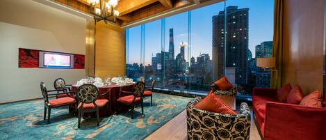 A hotel room with a city view, a dining area with a table set, and a seating area with patterned chairs and sofas.