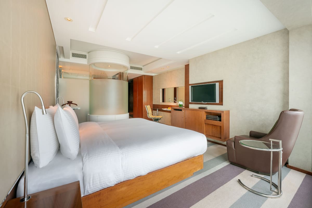 A modern hotel room with a large bed, a desk, a chair, a TV, and a shower area.