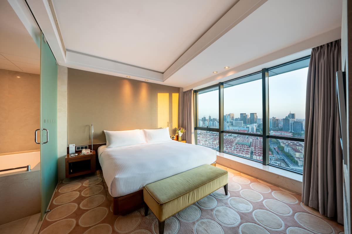 A modern hotel room with a large bed, a bench, a nightstand, and a view of the city through large windows.