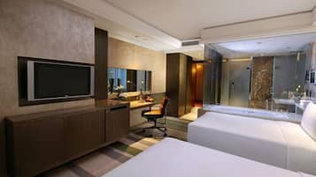 A modern hotel room with a large bed, a desk with a chair, a television, and a bathroom with a glass shower.
