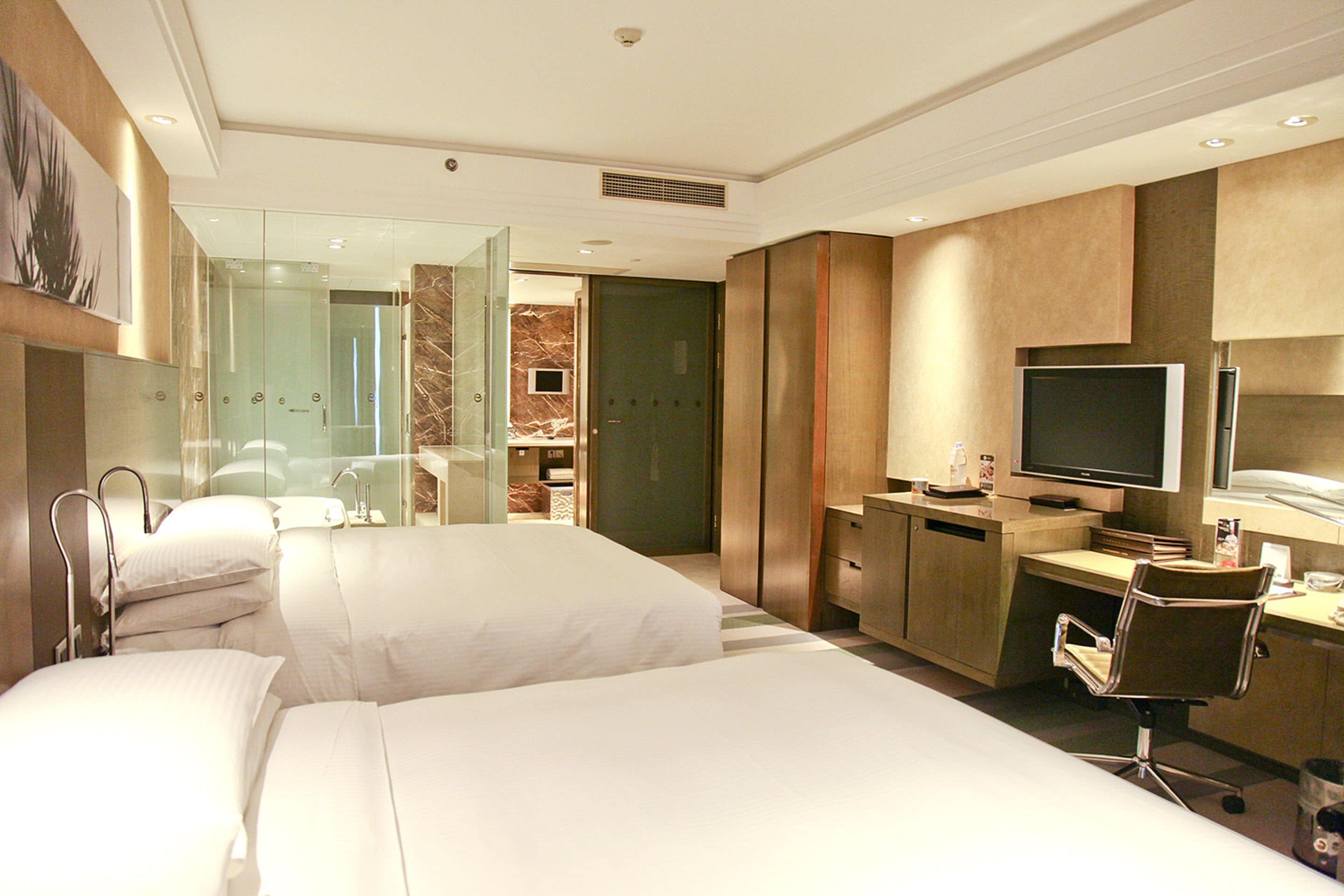 Habitación de hotel con dos camas, un escritorio con computadora, televisión y baño con cabina de ducha de vidrio.