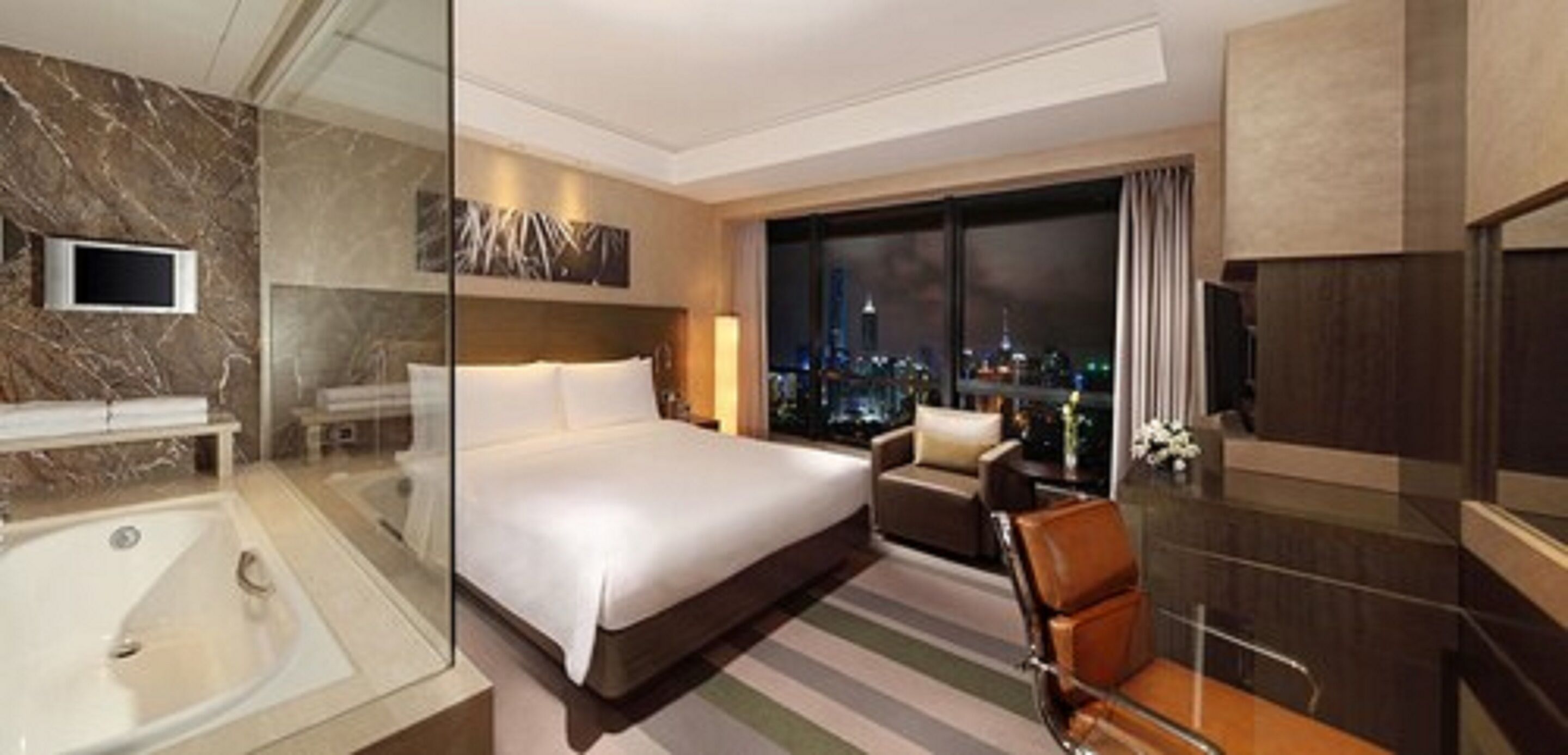 A modern hotel room with a large bed, a glass-enclosed shower, a city view, and a dining area with a view.