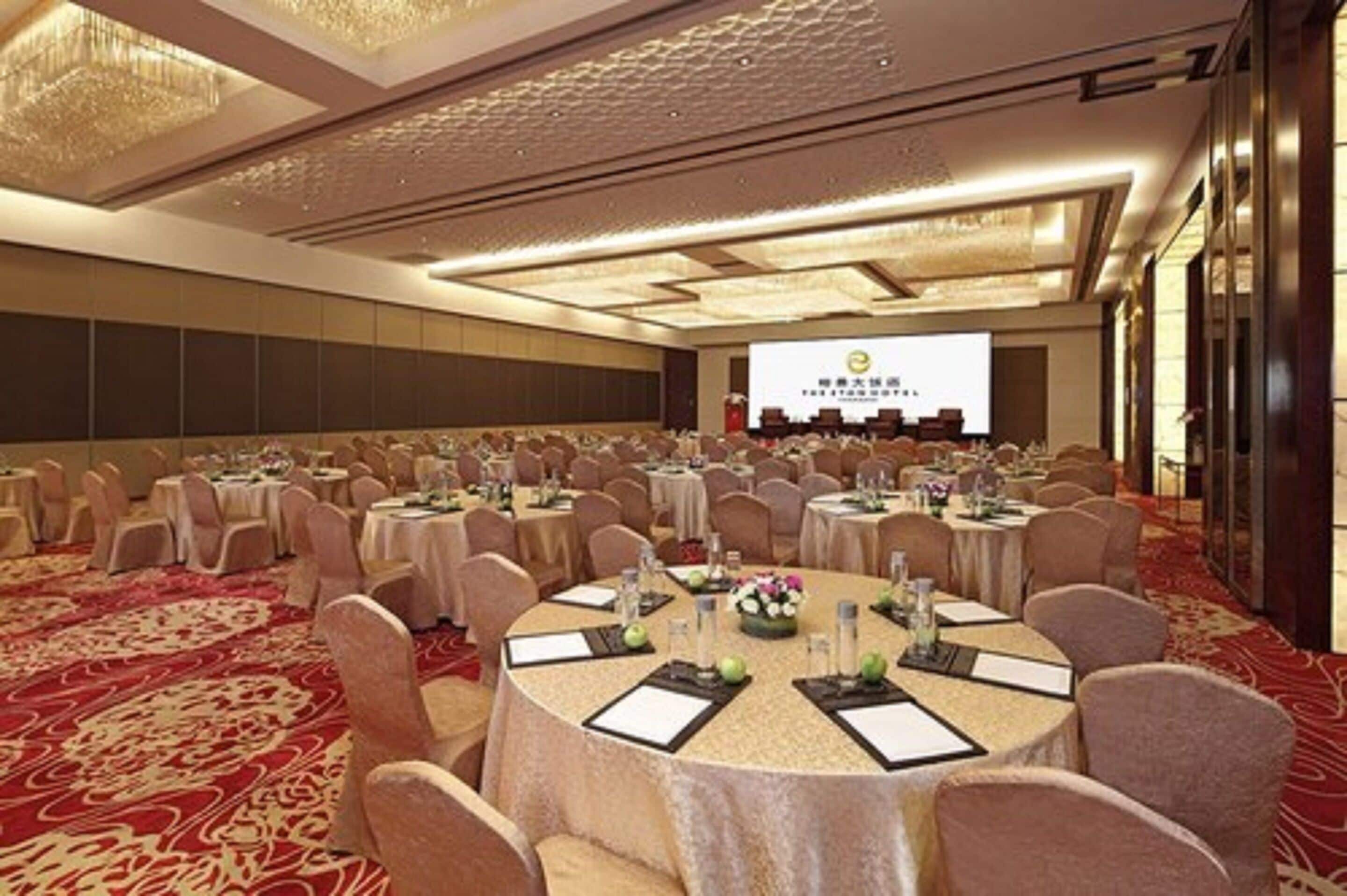 A banquet hall with round tables, chairs, and a stage with a presentation screen.