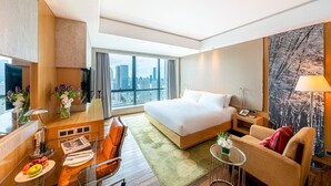 A hotel room with a large bed, a sofa, a desk with a lamp, a small table with flowers, and a city view through the window. - The Eton Hotel (Shanghai)