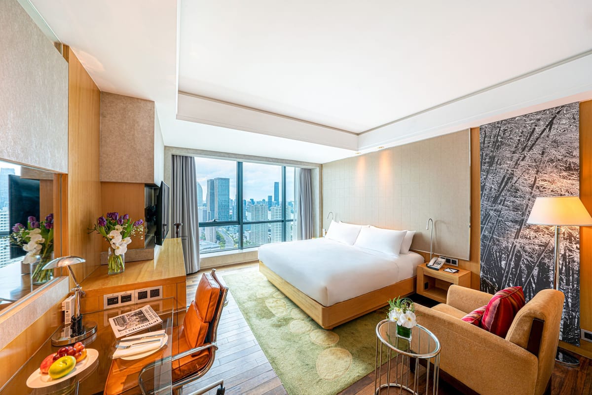 A hotel room with a large bed, a sofa, a desk with a lamp, a small table with flowers, and a city view through the window.