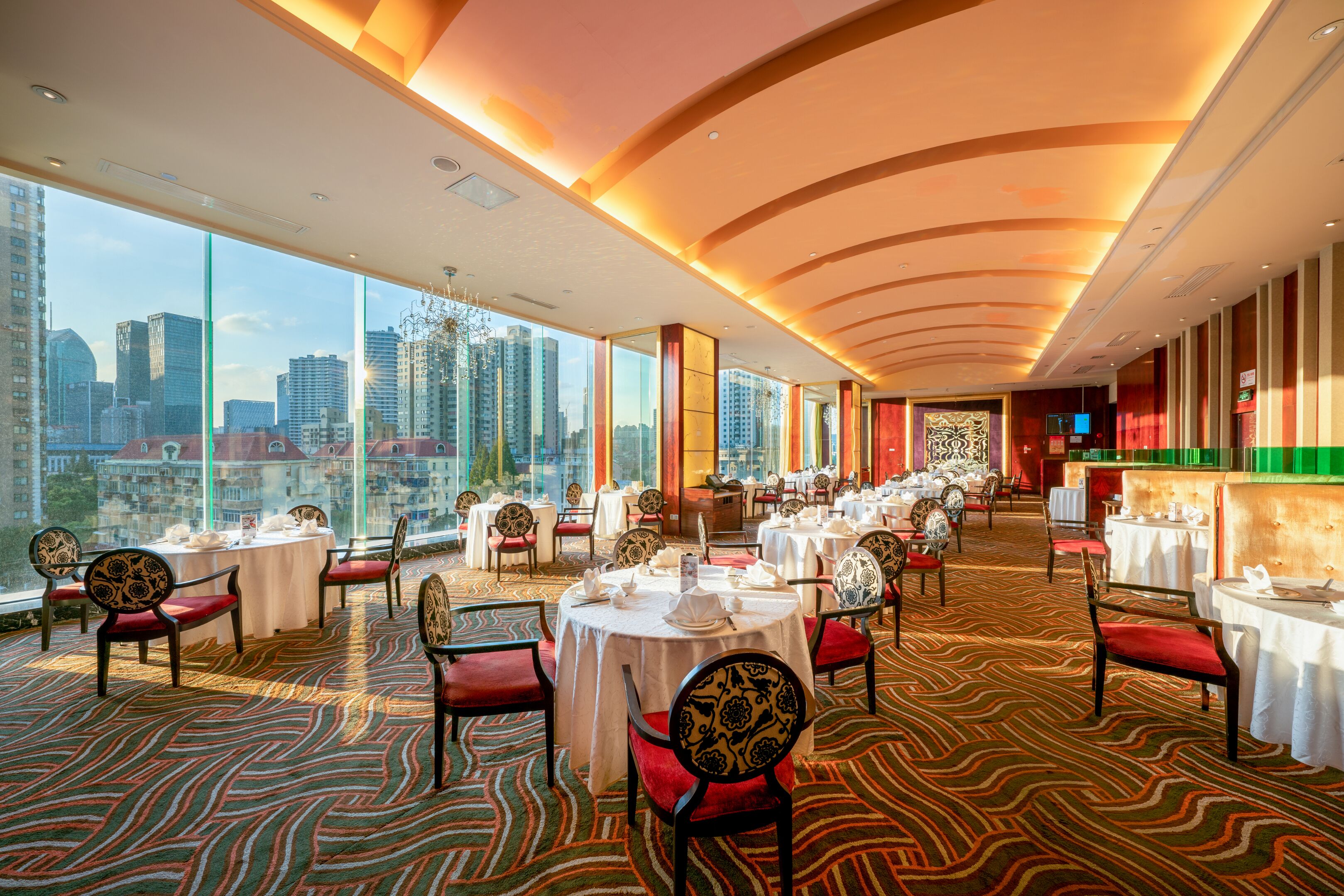 A spacious restaurant with high ceilings and large windows offering city views. Tables are neatly arranged with chairs, and the floor features a patterned carpet.