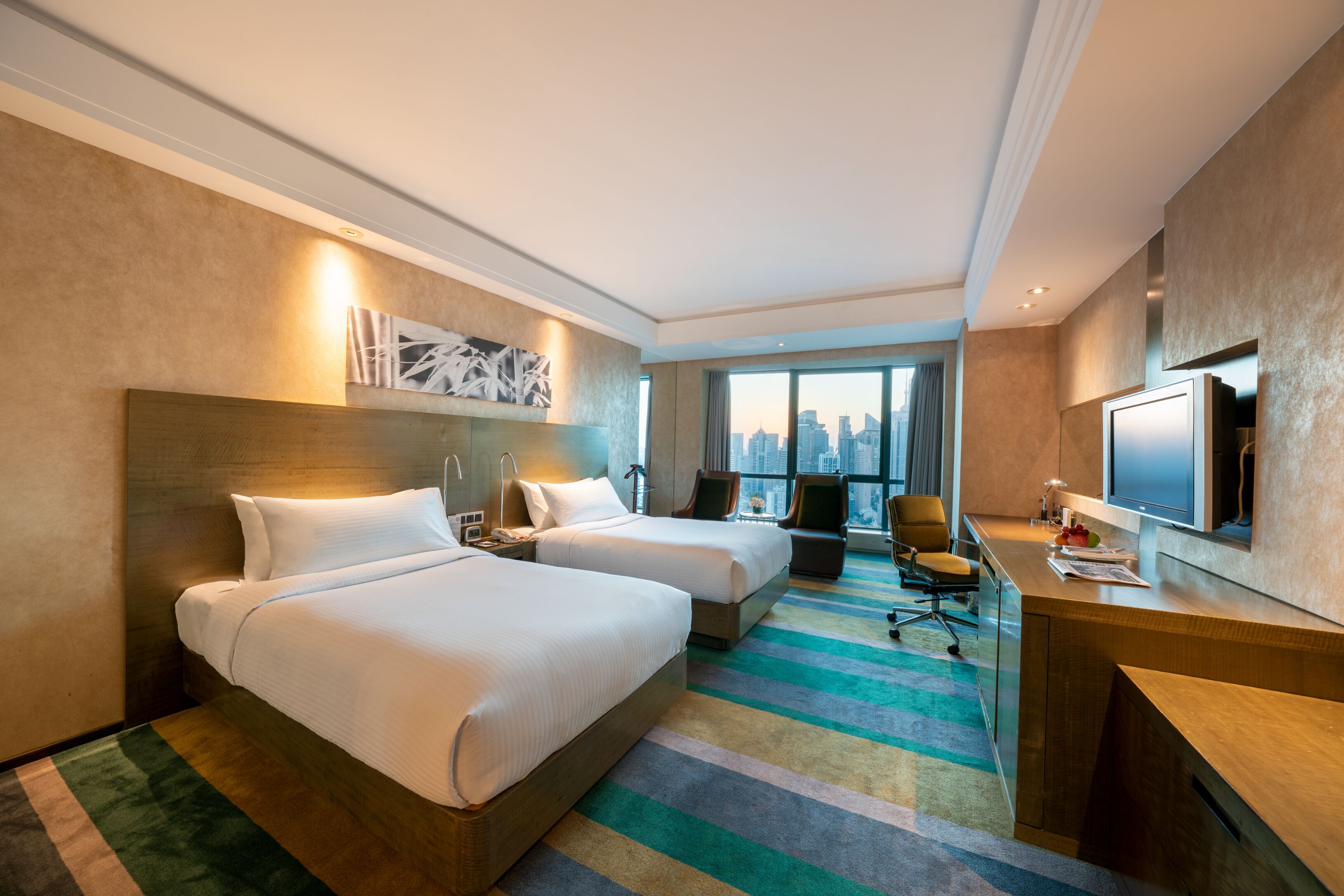A hotel room with two beds, a desk, a TV, and a large window with a city view.