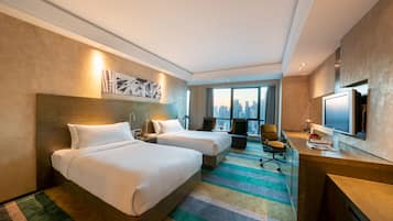 A hotel room with two beds, a desk, a TV, and a large window with a city view.