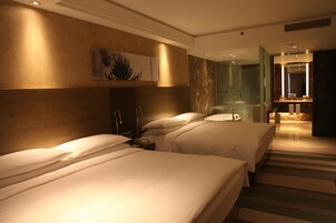 A hotel room with two beds, a bathroom with a glass shower, and a hallway leading to a dining area.