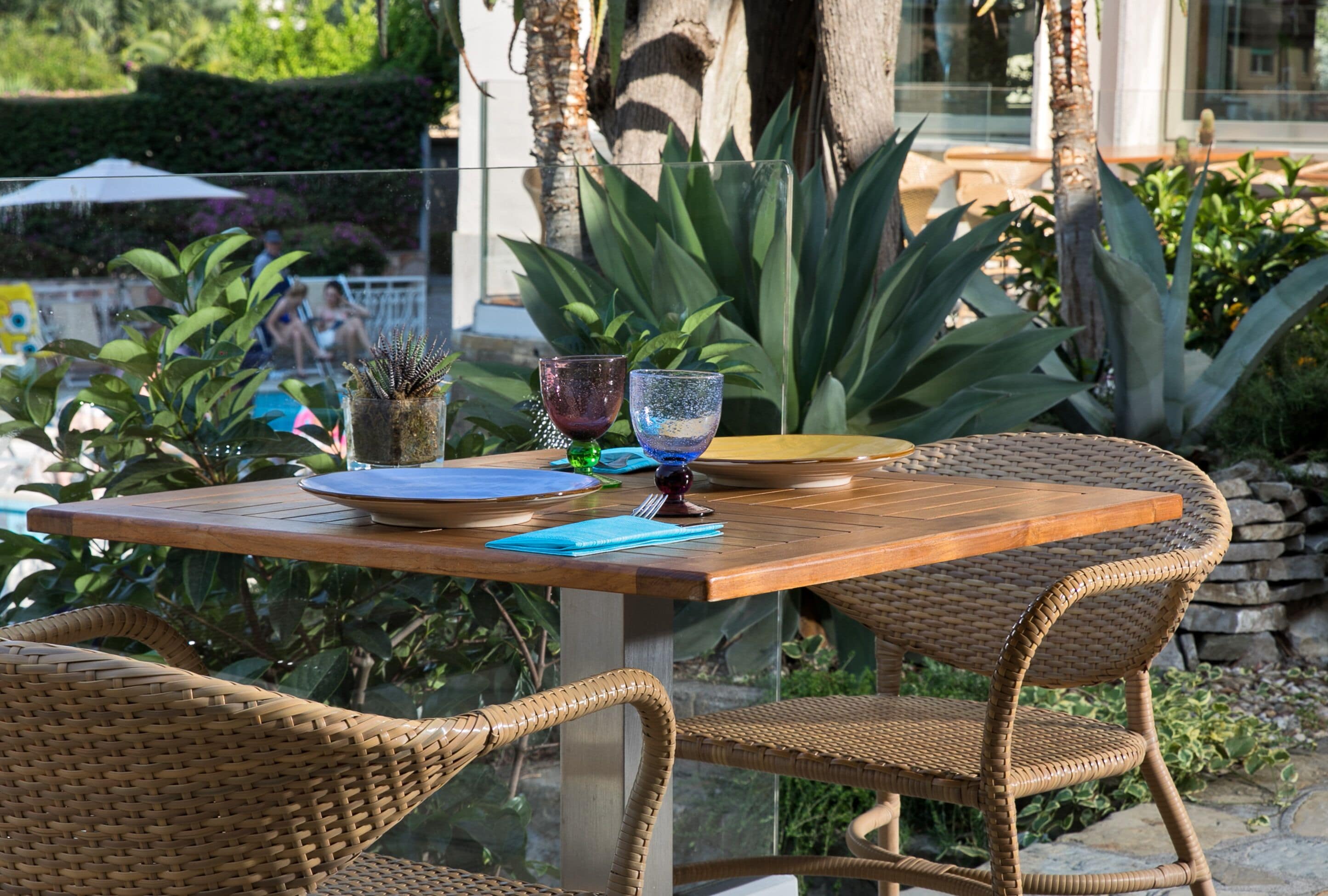 A wooden table with glass top, surrounded by wicker chairs, in an outdoor setting with plants and a stone wall.