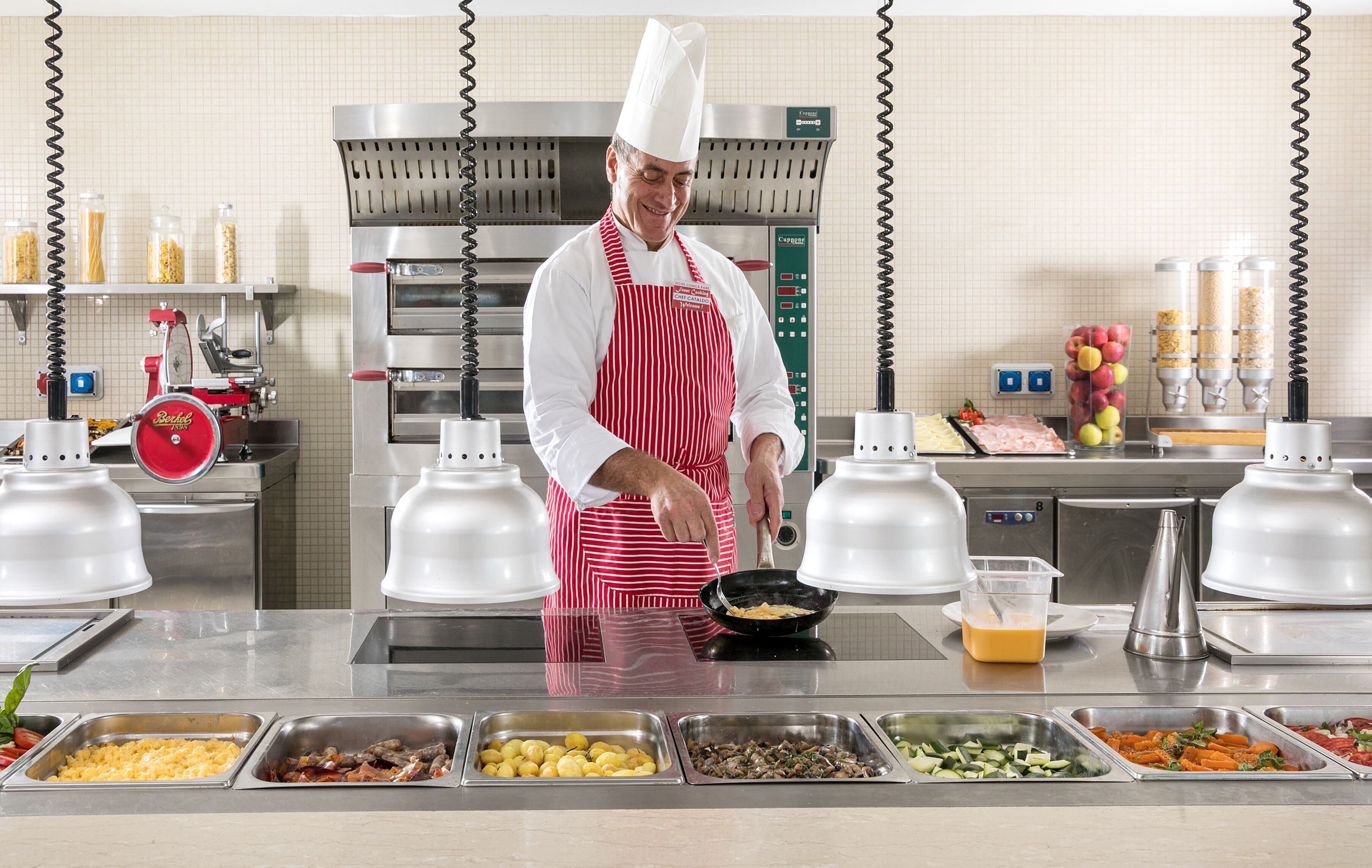 Um chefe de cozinha numa cozinha com várias estações de comida, um forno industrial e diversos recipientes de alimentos.