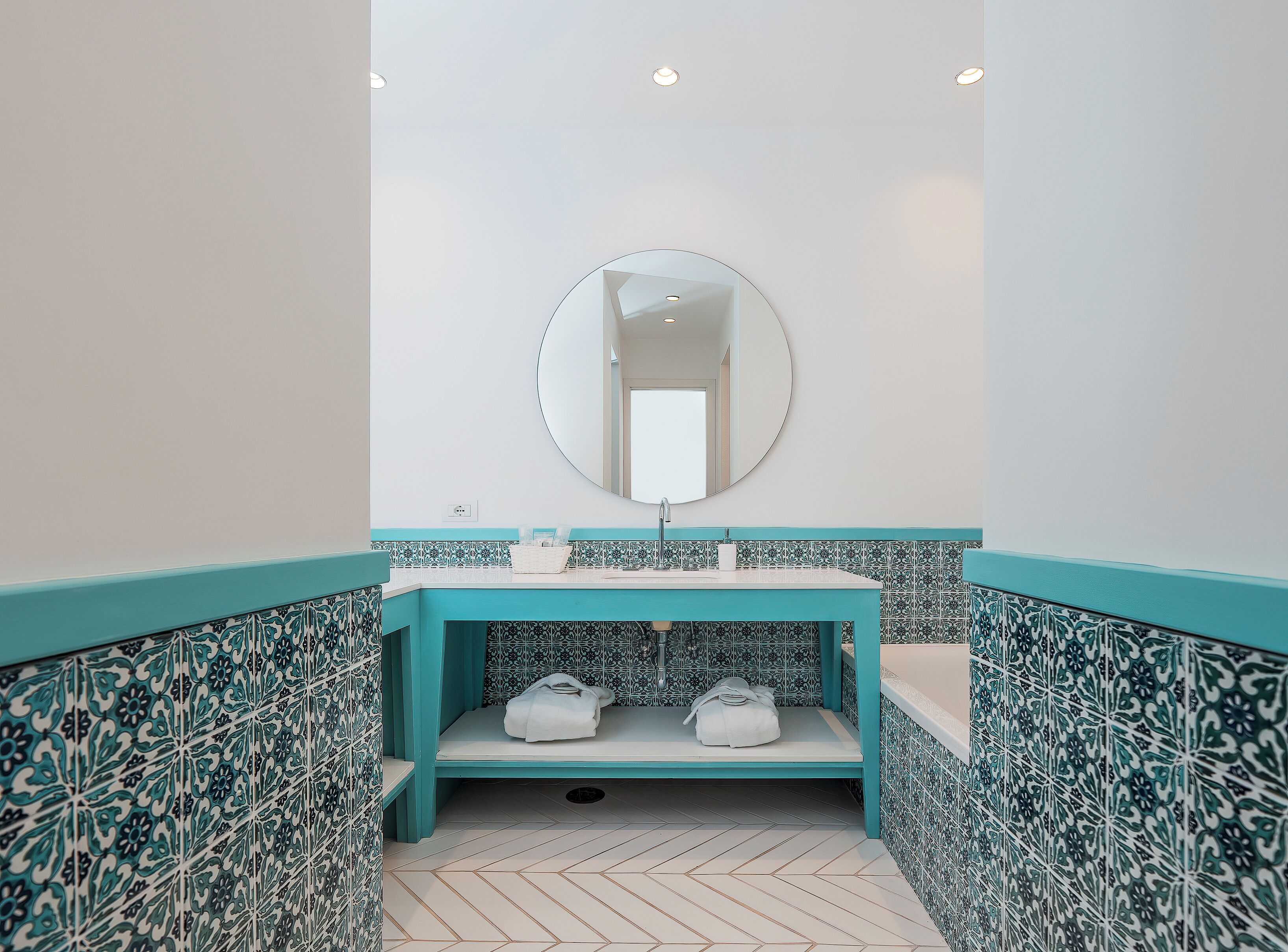 A bathroom with a white sink, a round mirror, and a bench with towels.