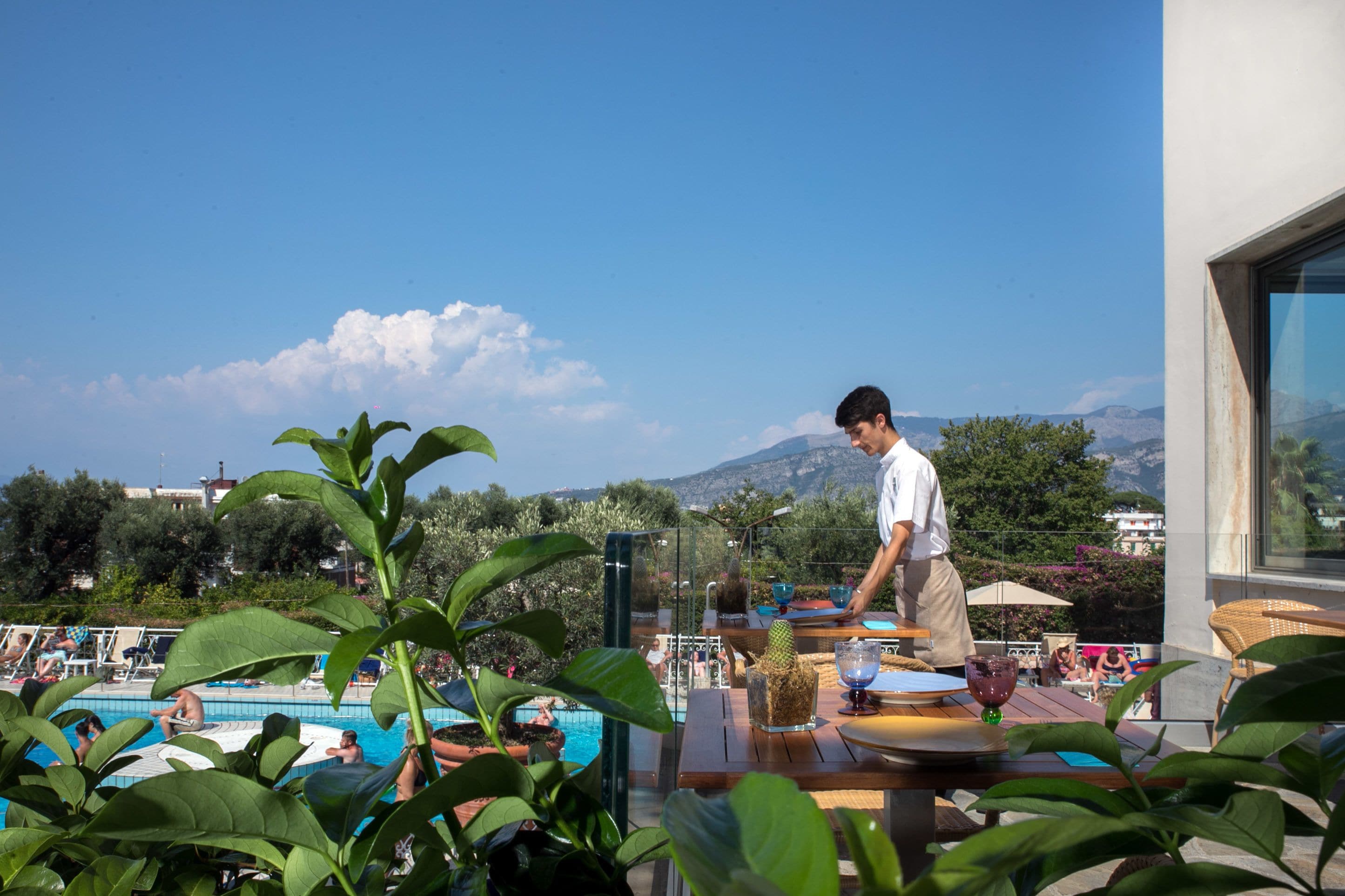 A poolside area with a view of mountains, a dining table with a glass of wine, and a person serving.