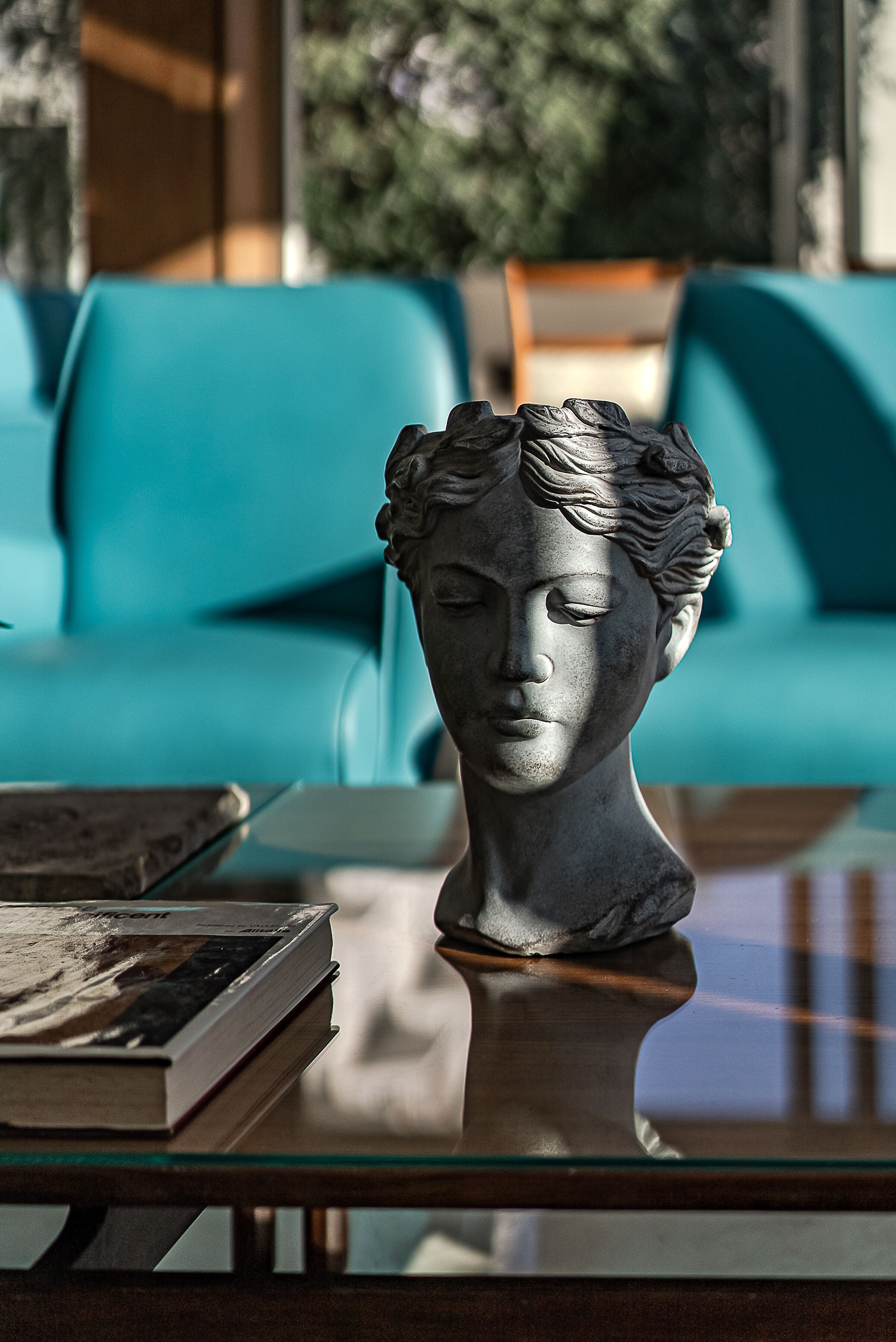 A bust sculpture on a glass table with a book beside it, in a room with teal sofas and a window.