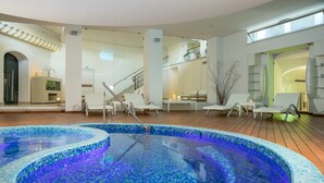 Piscina interior moderna con mosaicos, rodeada de sillones de descanso y una terraza de madera.