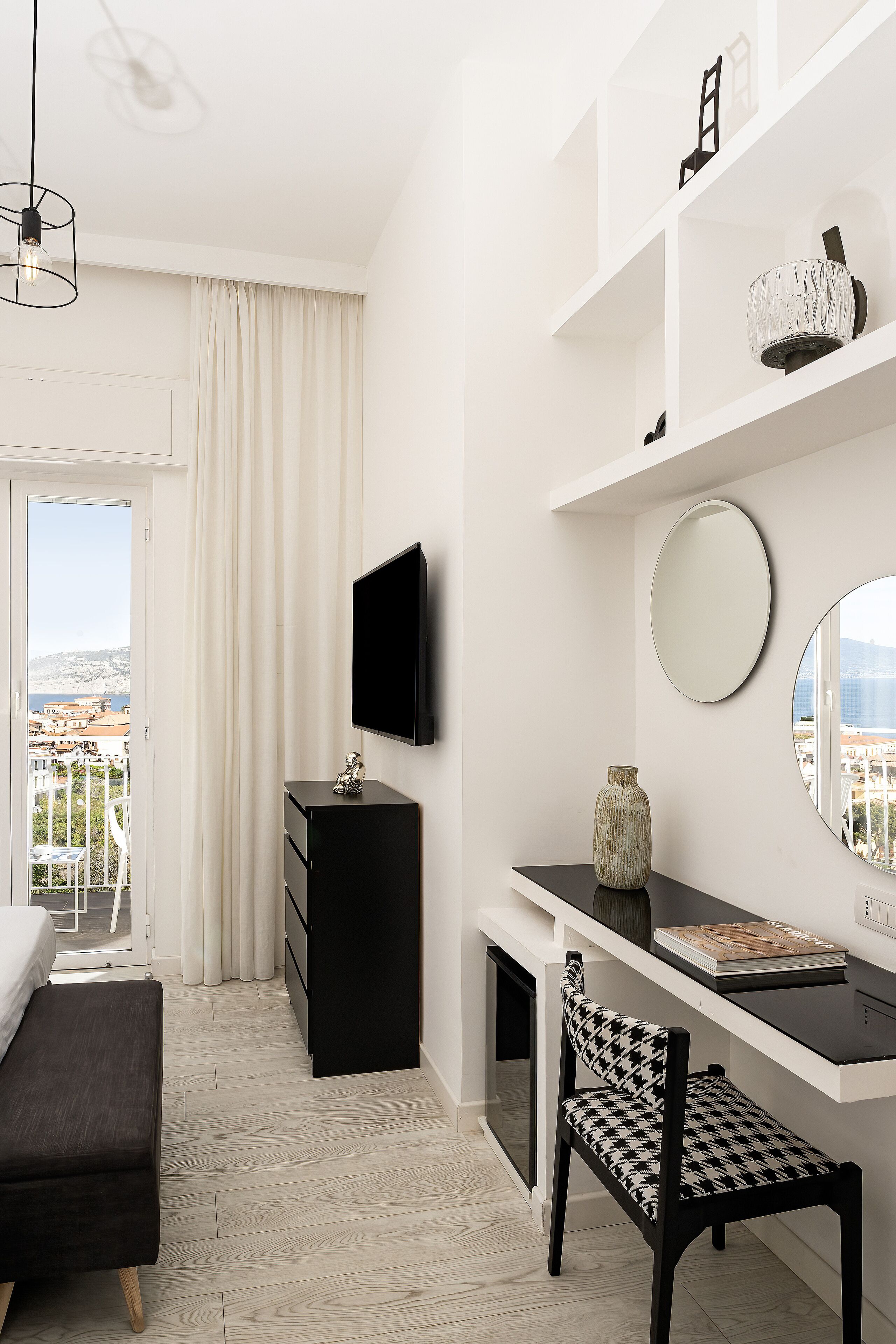 A modern hotel room with a balcony, a flat-screen TV, a round mirror, a black desk, and a houndstooth patterned chair.