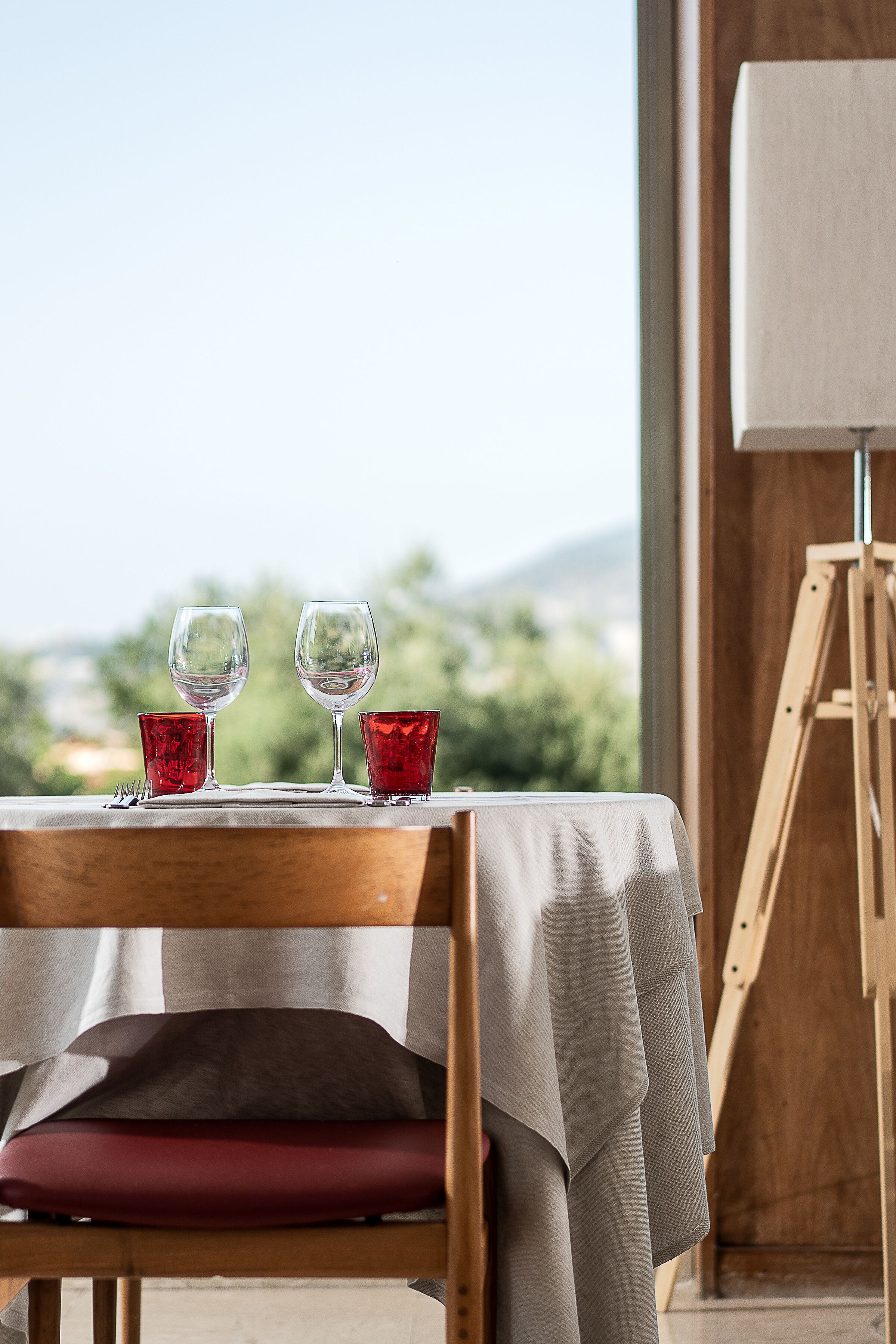 Une table à manger avec deux verres à vin et deux verres rouges, un chevalet en bois, et une vue sur la verdure et les montagnes.