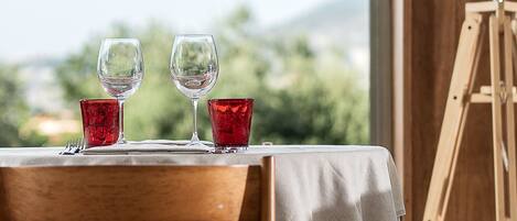 Une table à manger avec deux verres à vin et deux verres rouges, un chevalet en bois, et une vue sur la verdure et les montagnes.