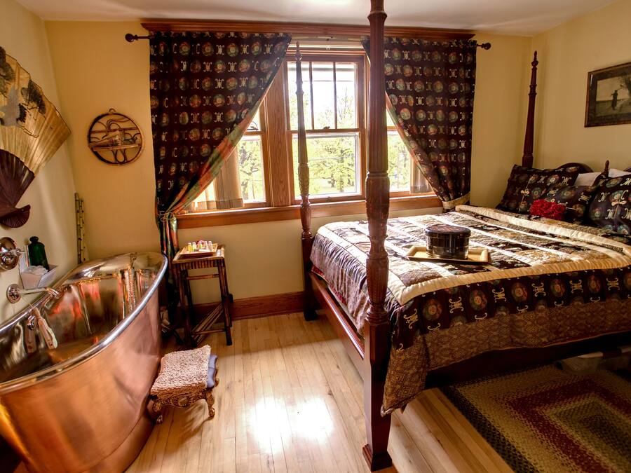 Un dormitorio tradicional con cama con dosel, piso de madera y un ventanal con cortinas.