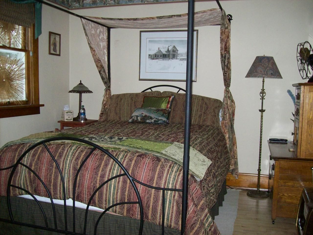 A bedroom with a four-poster bed, a bedside table with a lamp, a picture on the wall, and a window with curtains.
