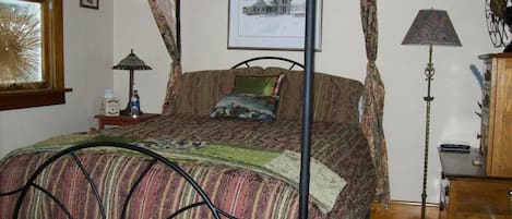 A bedroom with a four-poster bed, a bedside table with a lamp, a picture on the wall, and a window with curtains.