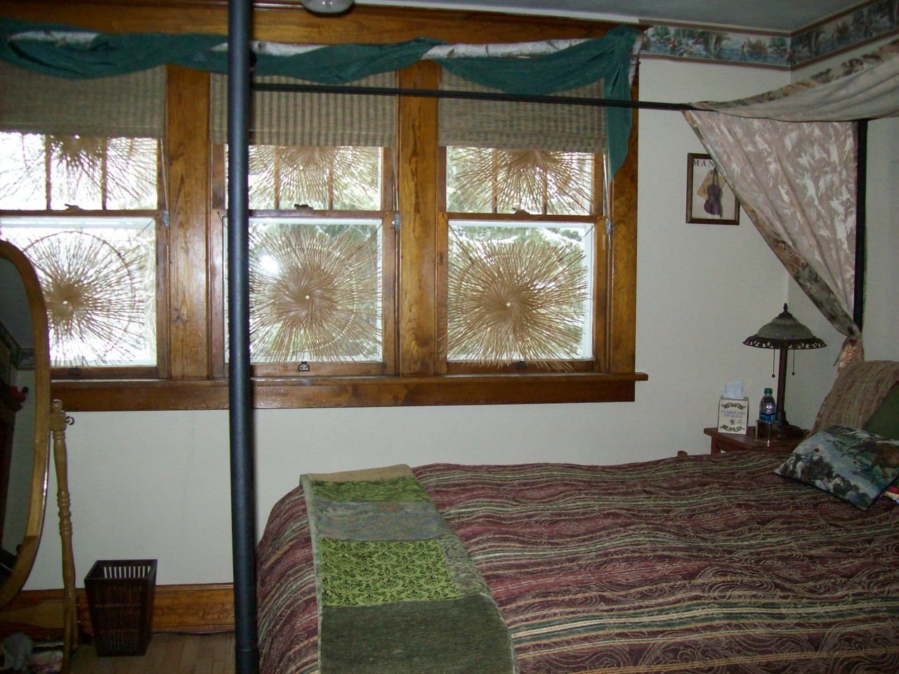 Un dormitorio con un ventanal grande, una cama con cubrecama estampado, una mesita de noche con lámpara y un espejo en la pared.